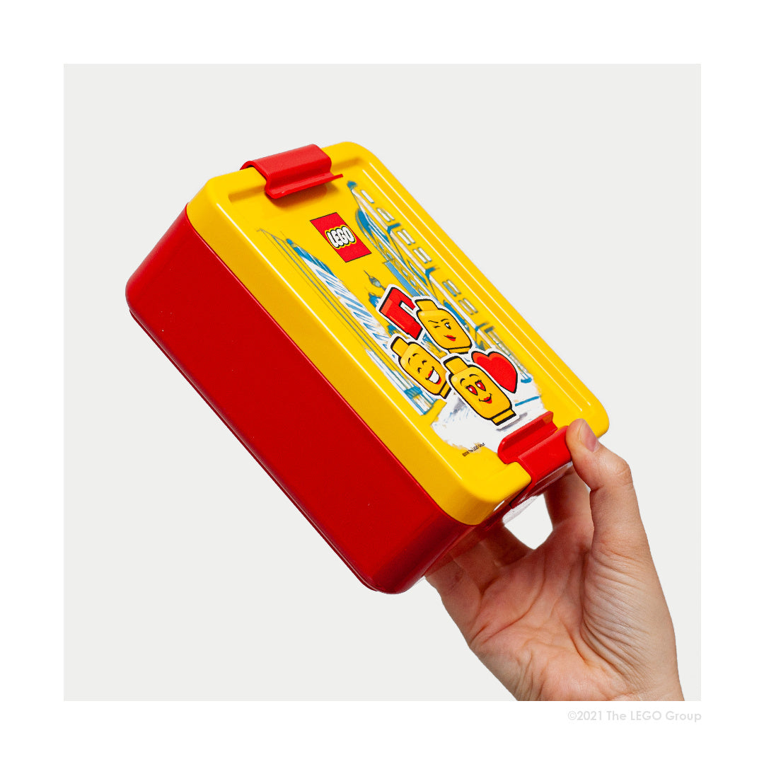 Lunchbox LEGO dla dziewczynki w czerwono-żółtych kolorach z motywem klocków LEGO trzymany w dłoni