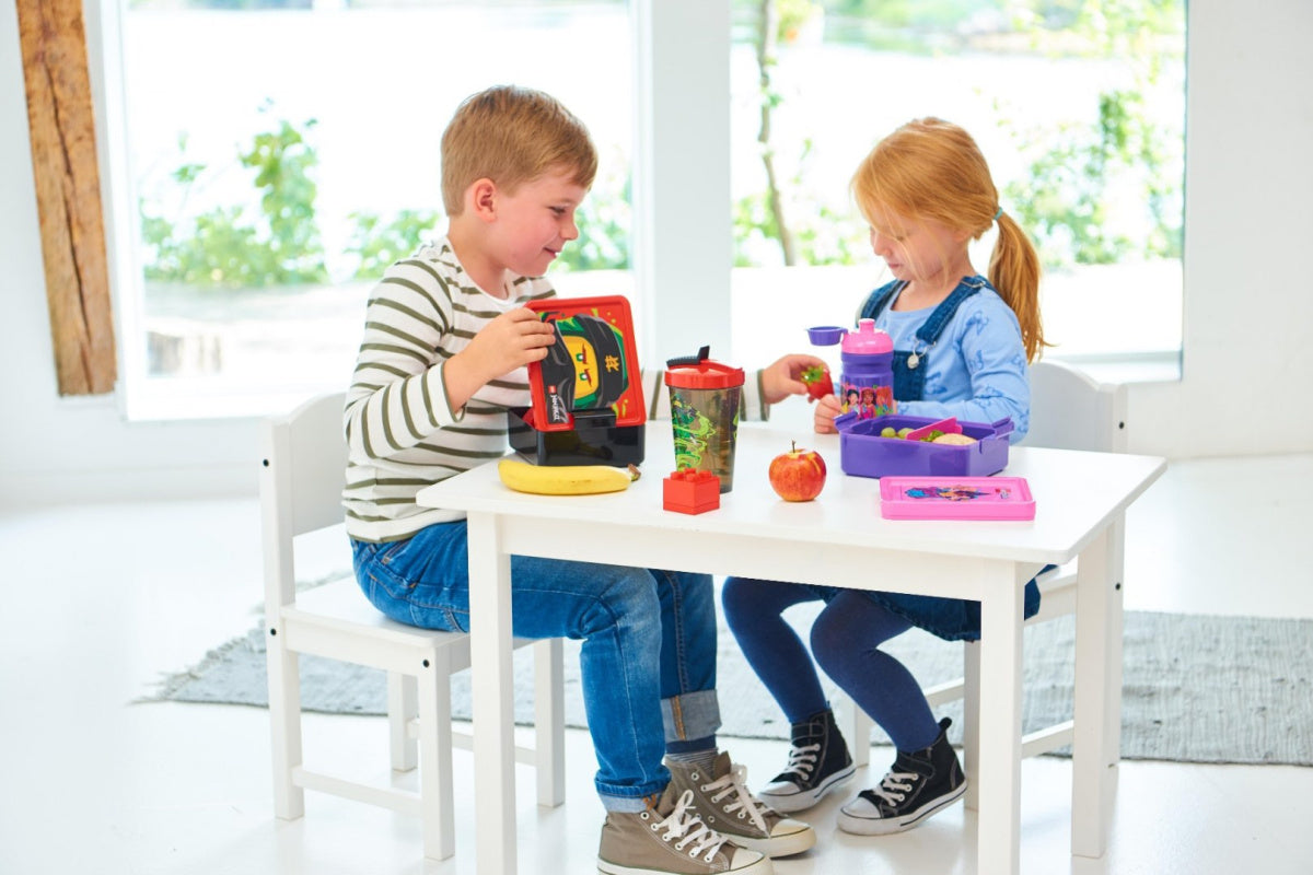 Chłopiec i dziewczynka jedzący posiłek z lunchbox LEGO City przy białym stole w jasnym pokoju
