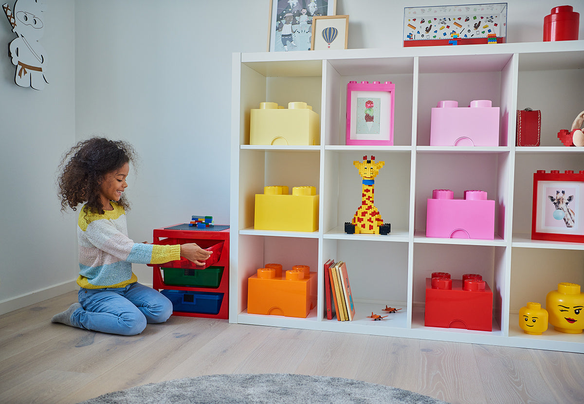 Dziecko bawiące się kolorowym pojemnikiem LEGO na klocki obok półki z innymi pojemnikami LEGO na klocki