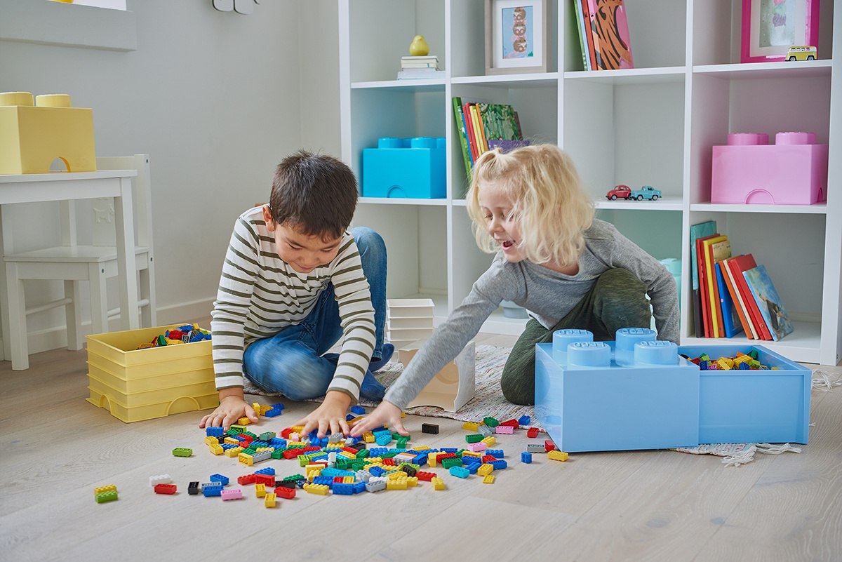 Dzieci bawiące się kolorowymi klockami LEGO na podłodze obok niebieskiego i żółtego pojemnika LEGO na klocki