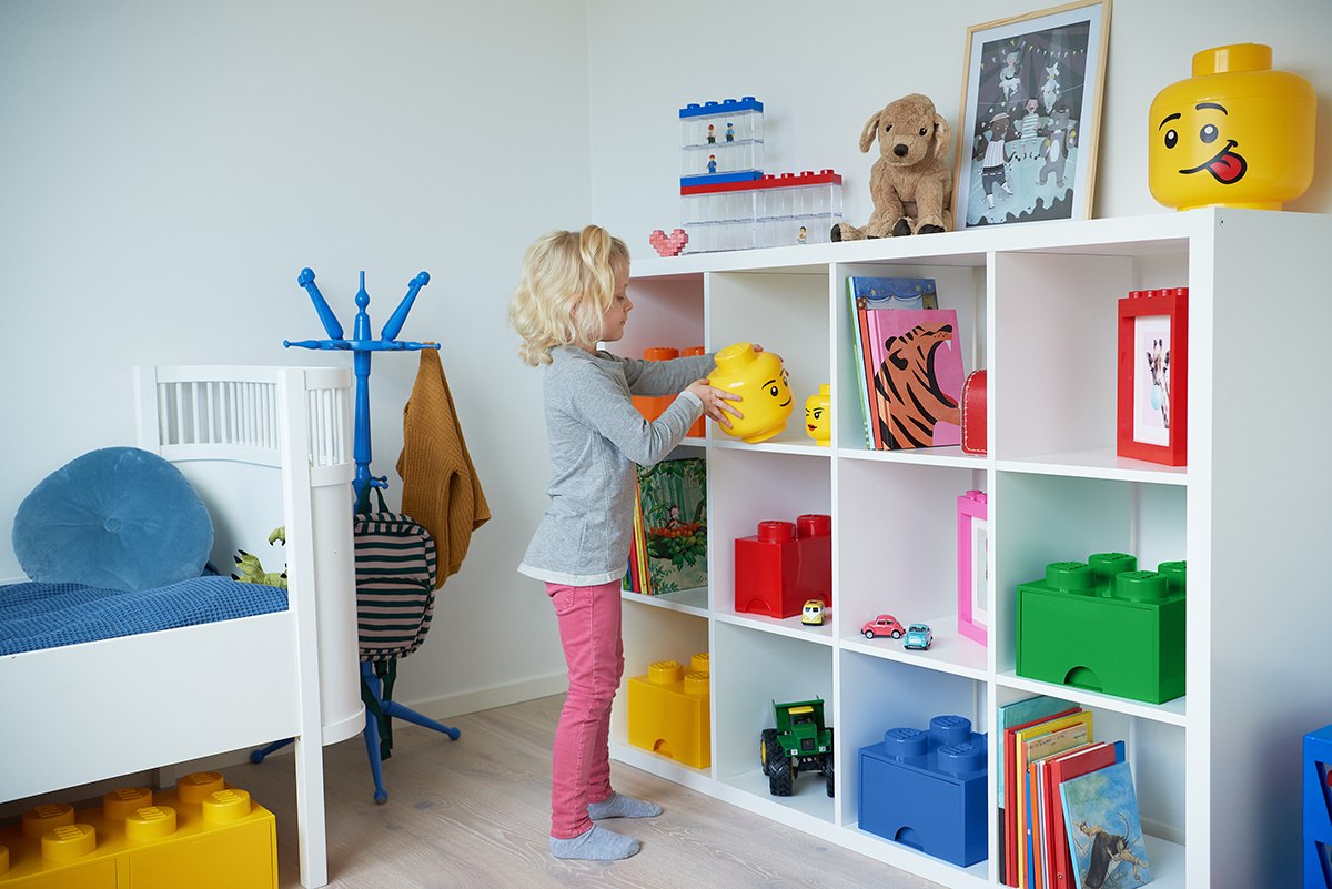 Dziecko trzymające żółty pojemnik LEGO na klocki na białej półce w pokoju dziecięcym