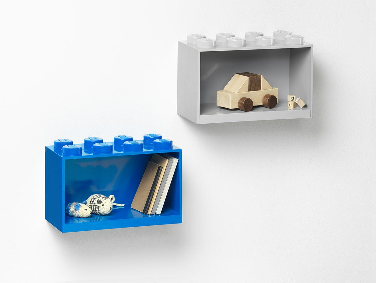 Biała półka LEGO w kształcie klocka z drewnianym samochodzikiem na białej ścianie obok niebieskiej półki z książkami i dekoracjami