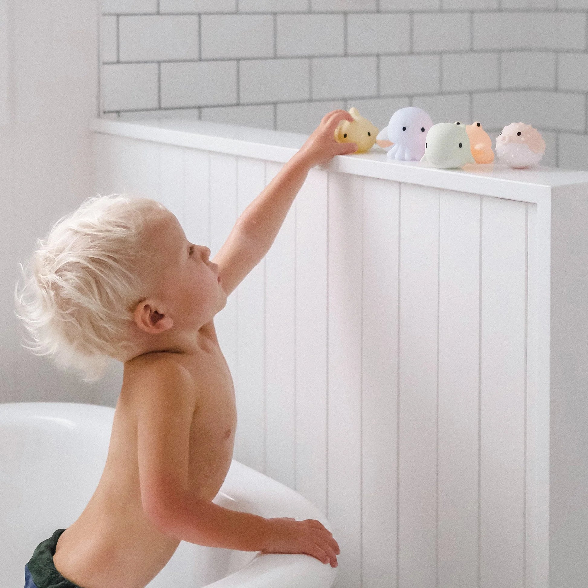 Dziecko sięgające po zabawki do kąpieli dla dzieci SUNNY LIFE na wannie w białej łazience